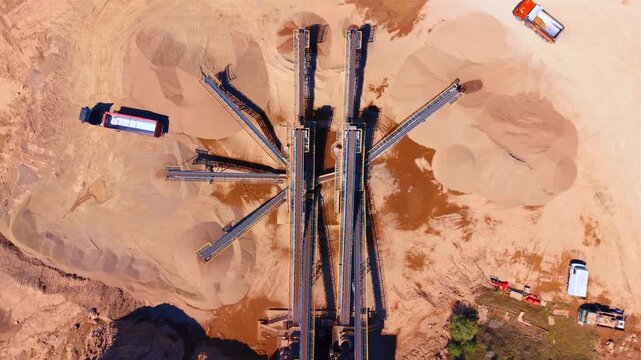 Aerial top down view of industrial conveyor system at a sand quarry. Mechanical conveyor belts extend over mounds of sorted gravel and dry soil.