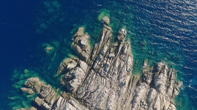 Vertical aerial footage of sculpted granite formations extending into the transparent Mediterranean Sea at Capo Testa, Sardinia, surrounded by vivid blue and turquoise water.
