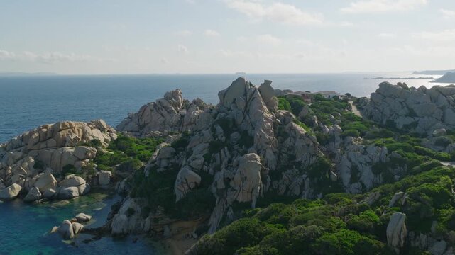 Cinematic aerial flight past towering granite formations and lush Mediterranean vegetation along the rugged turquoise coast of Capo Testa in northern Sardinia.