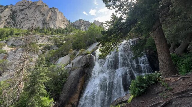 Wasatch mountain waterfall Utah, in a rocky mountain canyon, framed by pines and granite cliffs, with the water spilling over a broad rock face into a shaded pool below.