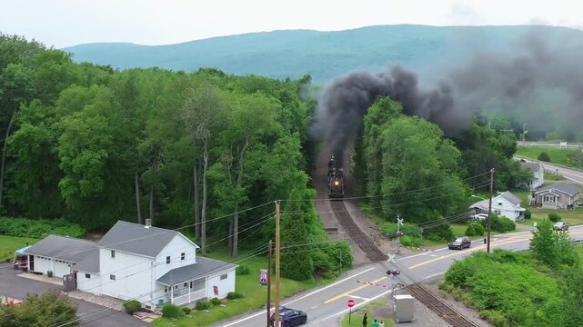 A steam train moves down the tracks while letting out black smoke. The train passes near houses in a green area. This happens in a rural place surrounded by trees.