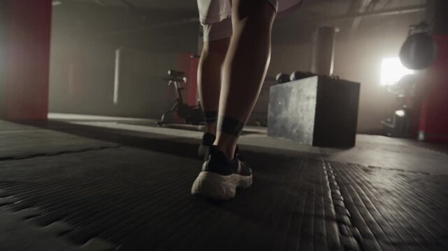 Young Caucasian woman walking in slow motion inside dark boxing gym, low angle shot focusing on legs and sneakers. Footage ideal for sports apparel commercials and fitness motivation videos