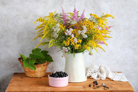 Wildflower bouquet in a vintage pitcher stands beside blackcurrants and scissors on a wooden table. Rustic summer still life concept for home decoration.