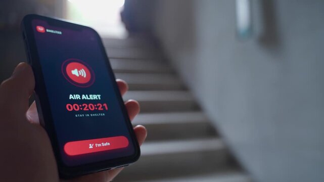 Hand holding smartphone with air raid alert app showing warning timer in stairwell 