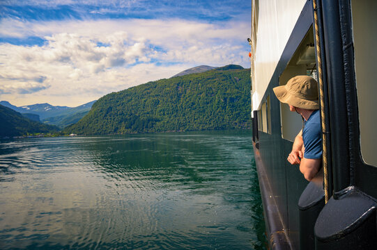 Passenger leans from a window aboard the Eidsdal-Linge ferry on Norddalsfjorden, surrounded by forested mountains, calm water, and dramatic clouds in More og Romsdal, Norway.