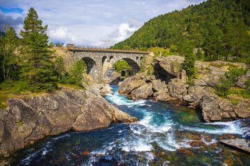Historic stone arch Romsdalen Bridge spans the rushing Rauma River in Norway's Romsdalen Valley, framed by rocks, forest, and green mountains. © Nick Fox