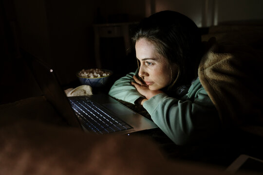 Woman with popcorn watching movie on laptop in dark while relaxing at home