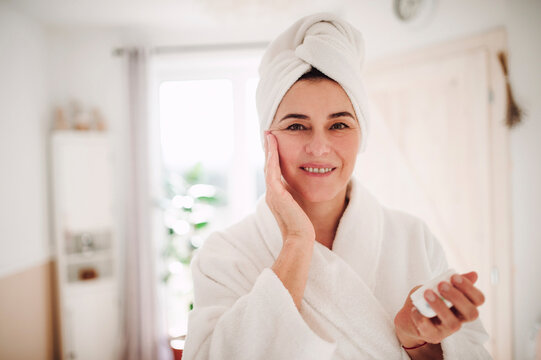Portrait of mature woman in a bathroom at home applying moisturizer