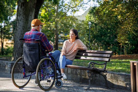 Smiling caregiver sitting on bench listening to retired man in wheelchair at park