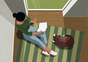 View from above of dog laying next to woman reading a book on a striped rug by a window at home