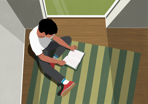 View from above of teenage boy reading book on striped book by window at home