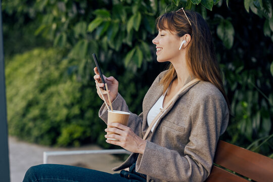 Woman uses smartphone and holds coffee cup on an outdoor bench
