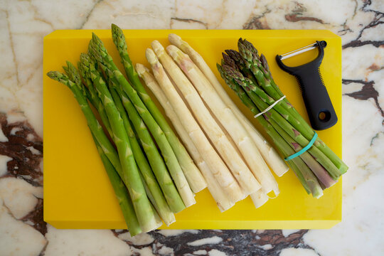 Still life view from above of green and white asparagus spears on yellow cutting board with peeler