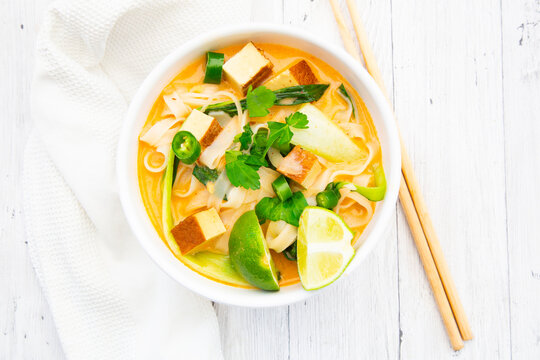 Red Thai curry soup with tofu and noodles sits on a wooden table