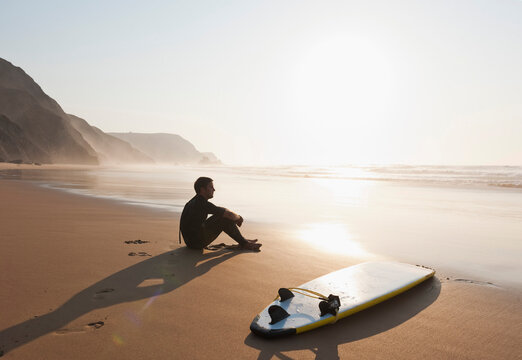 A male surfer watches the ocean from a sandy beach at sunset