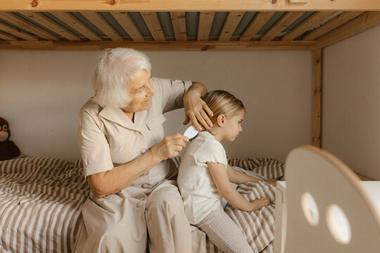 Grandmother combing hair of granddaughter sitting on bed at home