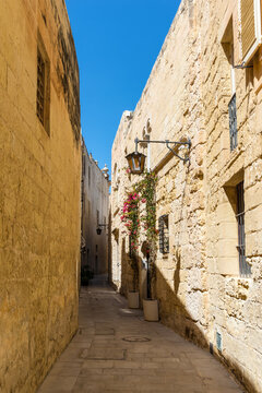 Narrow stone street lined with historic limestone buildings in the fortified city of Mdina, Malta. Authentic Mediterranean architecture with traditional lanterns and timeless old-world charm.