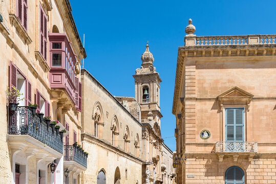 Charming street lined with elegant limestone buildings and a historic church bell tower in the fortified city of Mdina, Malta. Timeless Mediterranean architecture under a clear blue sky.