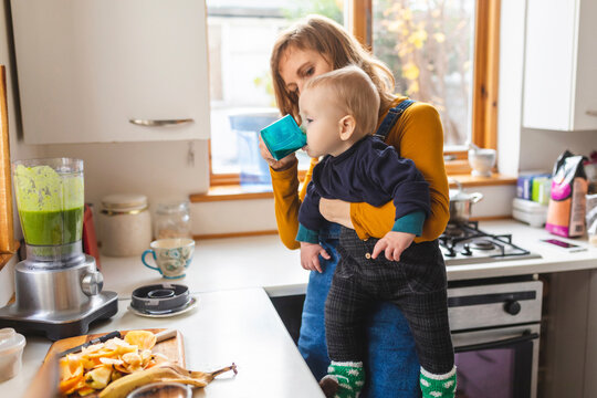 Woman in the kitchen preparing healthy smoothie and holding her baby son