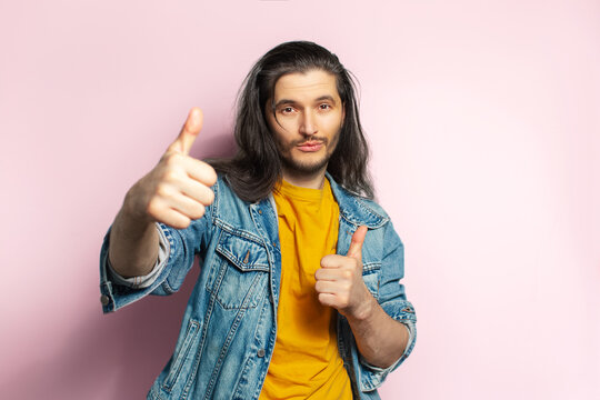 Young happy man with long hair showing thumbs up. Isolated on pastel pink background.