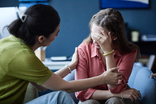 Young adult Caucasian woman covering face while female counselor comforting her during therapy session, expressing emotional distress and support. Ideal for mental health care campaigns