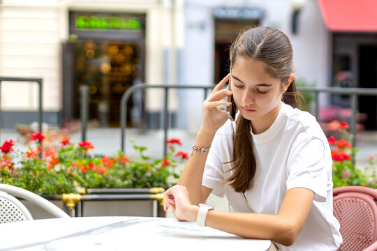 Young woman checking smartwatch during phone call at outdoor cafe. Young woman talks on a smartphone while checking her smartwatch at an outdoor cafe with red flowers and blurred city storefronts.