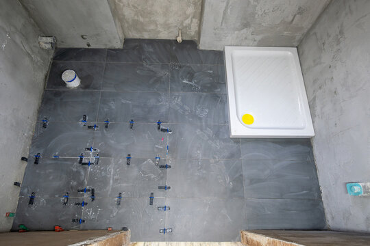 Bathroom floor being tiled with dark grey ceramic tiles and leveling system, installing a new white shower tray as part of a house renovation project