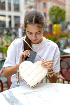 Young woman placing smartphone in heart-shaped handbag. Young woman places a smartphone into a quilted heart-shaped handbag while seated at an outdoor cafe.