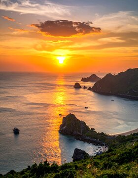 Aerial view of a coastline at sunset. The sun's light reflects on the water creating a golden hue. Green hills border the sea