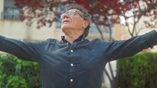 Man with glasses stretches arms wide and looks up amid apartment facade and red leaf tree outside building; joy gratitude freedom.
