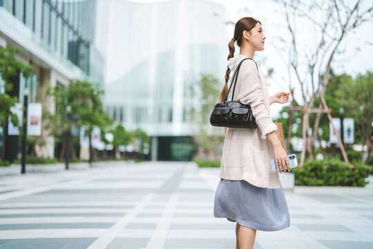 Young adult asian business woman side standing at outdoor on day wear sweater holding smartphone