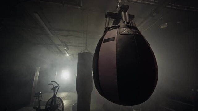 Heavy punching bag swinging in dark smoky boxing gym with exercise bike, captured in slow motion. Clip suitable for fitness brand marketing and athletic training promos
