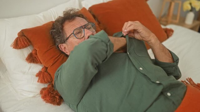Man in green shirt holds thermometer to forehead while lying on bed with orange pillows inside a building; fever illness worry.