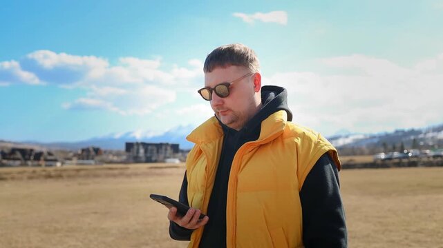 A young caucasian male tourist wearing a yellow vest and sunglasses stands outdoors, looking down at his smartphone. He is typing a message, checking a navigation map, or browsing the internet against