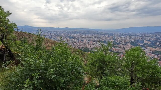 Tbilisi spreads beyond a forested hill in an elevated view, contrasting dense neighborhoods with mature trees and distant ridges to illustrate urban greenbelts, land management, and city planning.