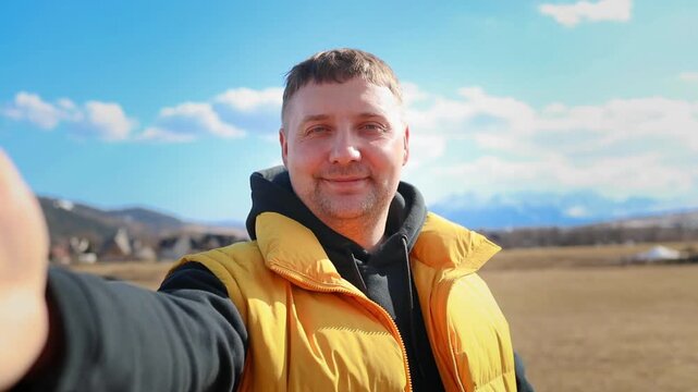 A cheerful young caucasian man in a yellow vest is holding a phone to record a selfie video or make a live stream call outdoors. He is smiling and waving his hand at the camera against a beautiful