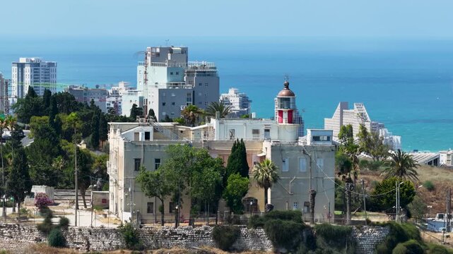High angle drone shot panning across the historic Stella Maris monastery lighthouse building on Mount Carmel, revealing the modern city highrises and Mediterranean shoreline in the background.