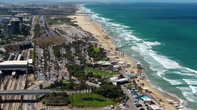 Elevated tracking view showcasing turquoise ocean waves crashing on a sandy beach adjacent to a bustling multi lane transit corridor and waterfront development.