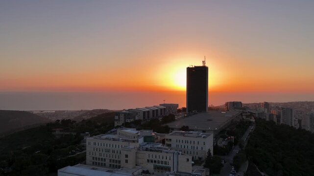 Drone tracking shot capturing a dramatic orange sunset behind a prominent university skyscraper on Mount Carmel overlooking the Mediterranean Sea.