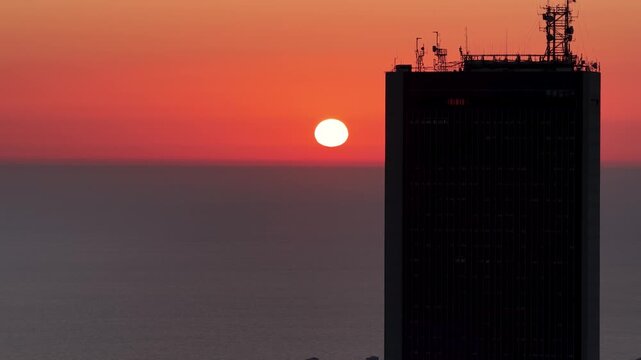A steady tracking shot capturing a dramatic orange sunset over the open sea as the sun dips behind the silhouette of a modern corporate highrise building.