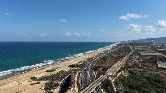 Drone flight capturing a modern passenger train moving on tracks parallel to vehicle traffic on a major coastal highway running alongside a sandy Mediterranean beach on a bright sunny day.