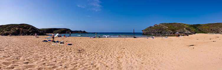 Fototapeta premium Wide panoramic view of a sunny sandy beach with rocky cliffs, calm blue sea and people relaxing, swimming and surfing on a warm summer holiday at the coast.