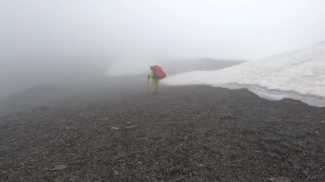 Female hiker crosses a foggy alpine snowfield in Georgia with a loaded backpack, navigating dark scree during a demanding high-altitude trek suited to adventure travel, endurance and outdoor safety.