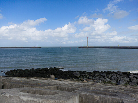 A calm harbor entrance with concrete breakwater and rocky shore, distant lighthouses and a solitary sailboat on the blue sea beneath a bright sky filled with scattered white clouds.