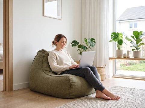 Relaxed Workspace: A woman comfortably uses a laptop while seated on a plush beanbag chair in a bright, airy room, creating a cozy and productive home office environment.