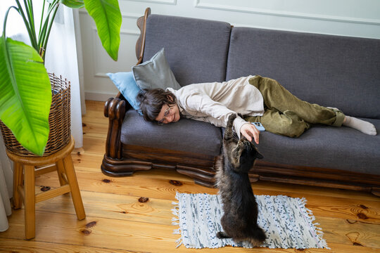 Cheerful woman playing with cat while fully relaxing on couch in room, happiness at home on weekend, lazy positive female resting on sofa enjoying fun moments with pet, self care and peaceful mood.