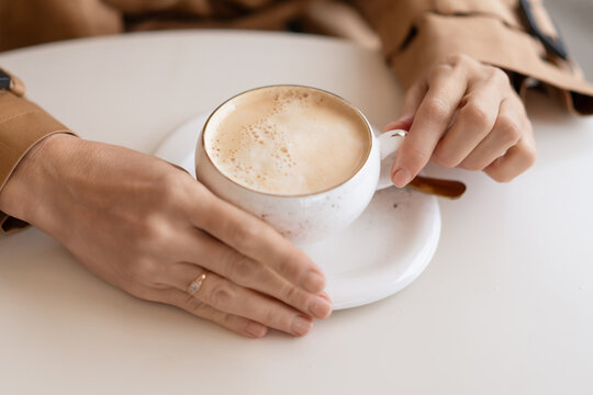 Morning cappuccino routine enjoyed by woman holding ceramic coffee cup at white cafe table