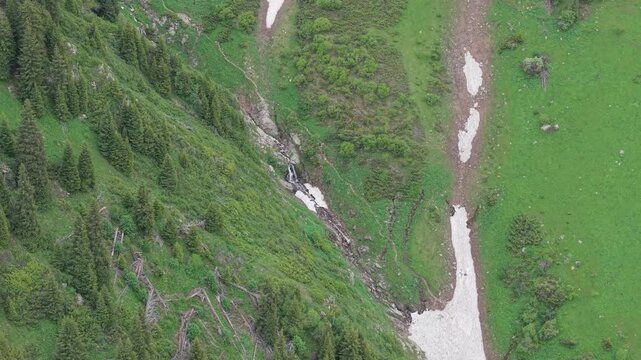 Aerial footage captures a cascading waterfall flowing down a lush green mountainside, surrounded by dense coniferous trees and patches of snow, showcasing the natural beauty of the landscape