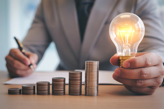 Business idea generation with light bulb stacks of coins and person writing.