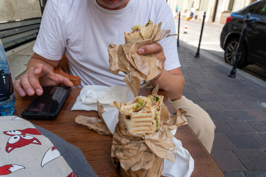 A casually dressed man holds a partially eaten grilled wrap filled with meat, lettuce, onion and sauce while sitting at an outdoor cafe table. A second wrap, smartphone and bottled water accompany the